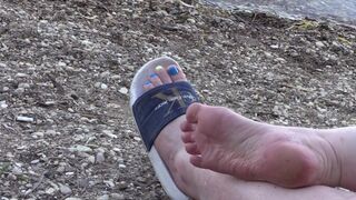 Legs with a Pedicure Sunbathe on the Beach. Foot Bizarre in Slippers